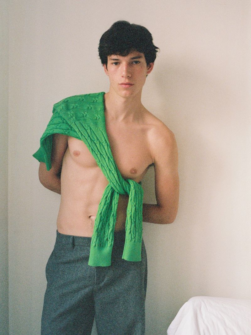 Shirtless male model wearing grey trousers with a bright green cable-knit sweater draped over his shoulder against a pale wall.