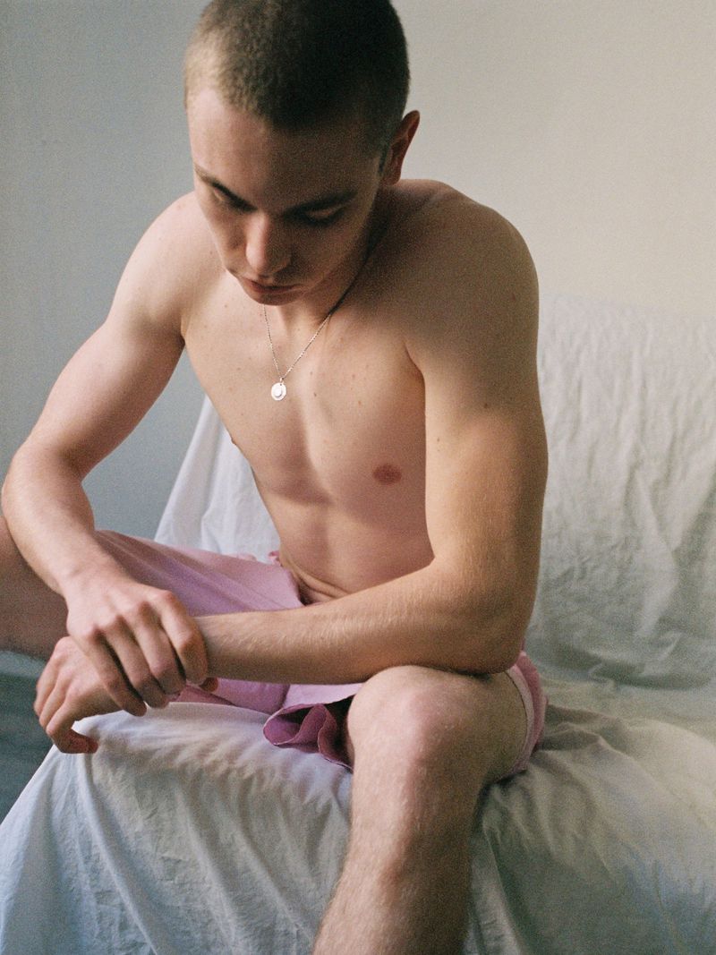 Shirtless young man with a buzz cut sitting in pink shorts and looking down, seated on a white draped chair.