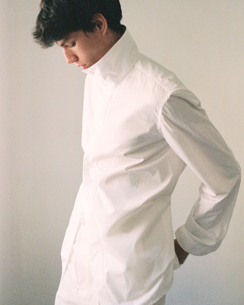 Male model wearing a crisp white button-down shirt with a structured high collar, looking down against a pale studio wall.