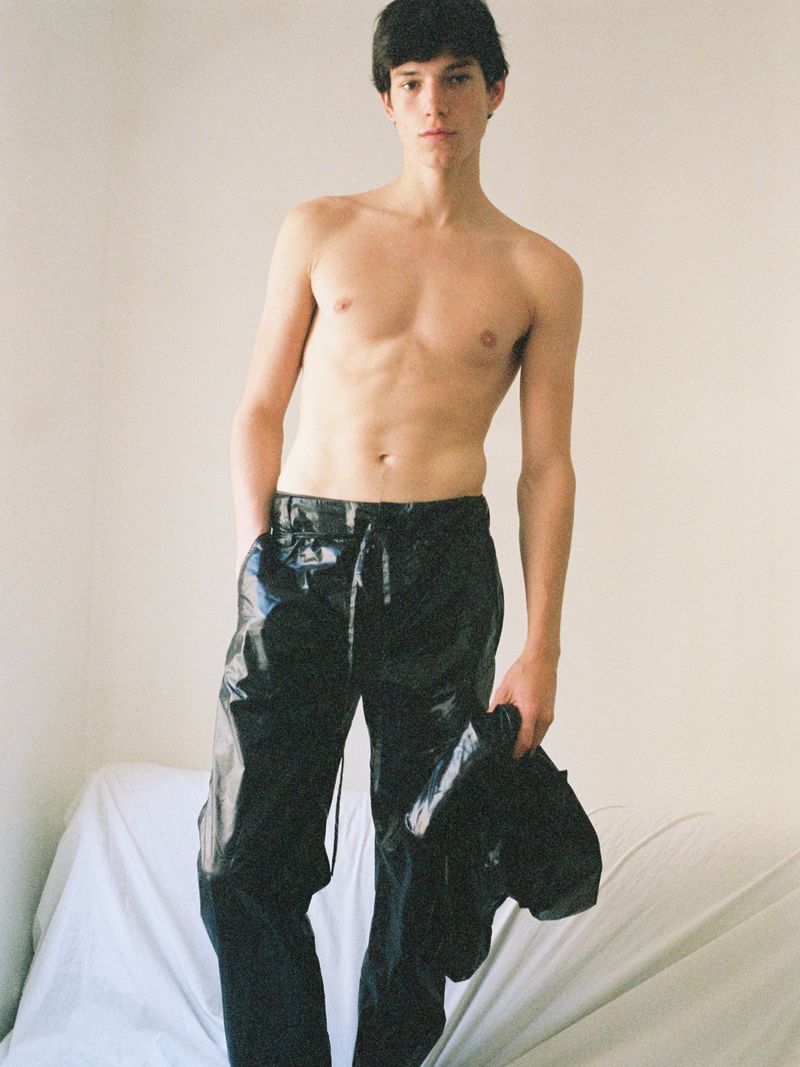 Shirtless male model wearing glossy black trousers and holding a black jacket against a white draped backdrop.