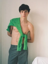 Shirtless male model wearing grey trousers with a bright green cable-knit sweater draped over his shoulder against a pale wall.