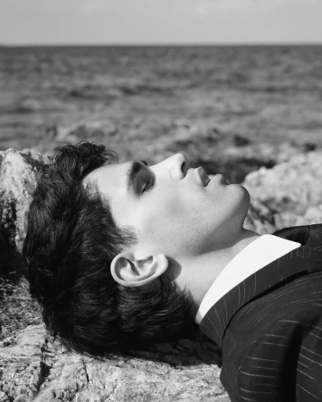 Monochrome portrait of a man lying on rocks by the ocean, eyes closed, wearing a suit jacket against a light blue sky.