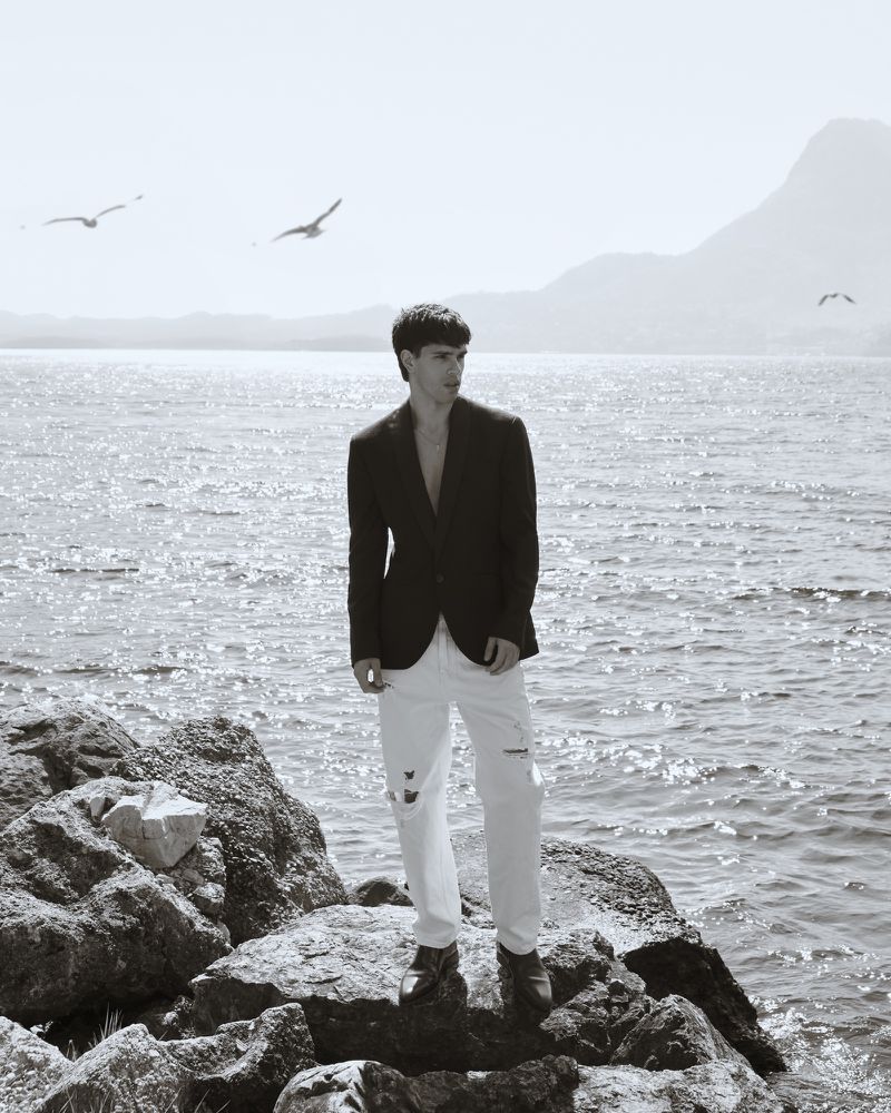 Monochrome portrait of a young adult man in a blazer standing on rocks by the sea with birds flying and a mountain in the background.