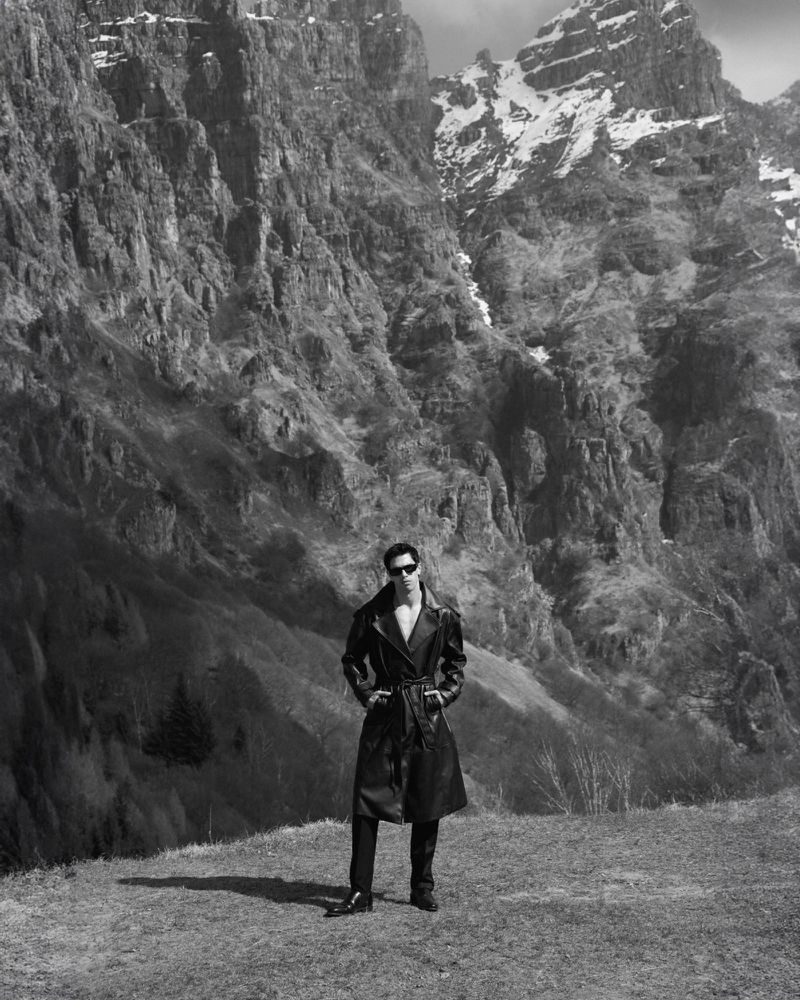 Model Denis Jovanovic standing in a black belted leather trench coat and sunglasses against towering rocky mountain cliffs.