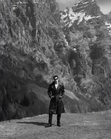 Model Denis Jovanovic standing in a black belted leather trench coat and sunglasses against towering rocky mountain cliffs.