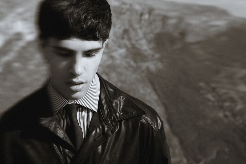 Monochrome portrait of an adult man with dark hair, wearing a striped shirt, tie, and leather jacket, set against a blurry mountain backdrop.
