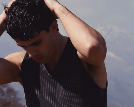 Young man with dark hair in a black ribbed tank top poses outdoors with a backdrop of distant mountains on a hazy day.