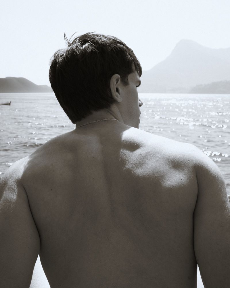 A shirtless man viewed from behind looking across sparkling sunlit open water toward distant mountains.