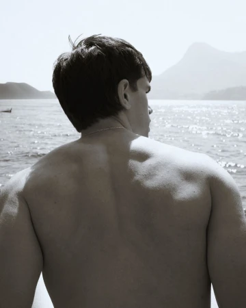 A shirtless man viewed from behind looking across sparkling sunlit open water toward distant mountains.