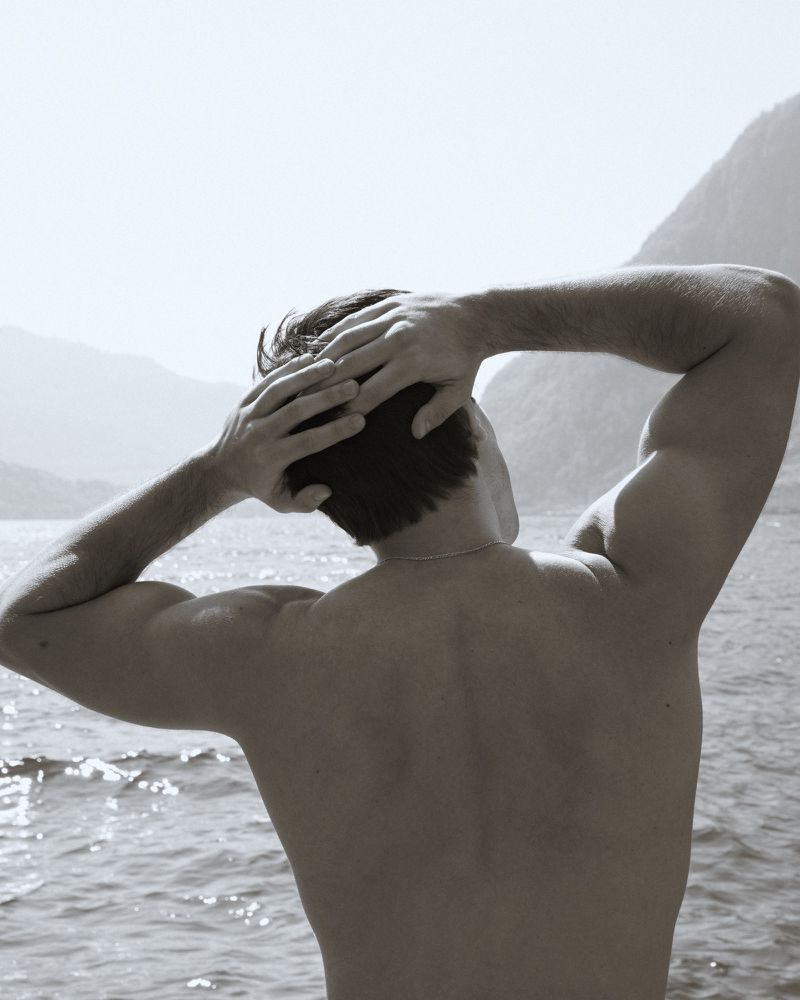 Back view of an adult male with his hands behind his head, standing outdoors near a lake with mountains in the background in monochrome.