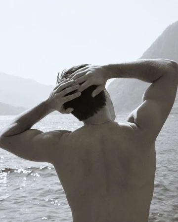 Back view of an adult male with his hands behind his head, standing outdoors near a lake with mountains in the background in monochrome.