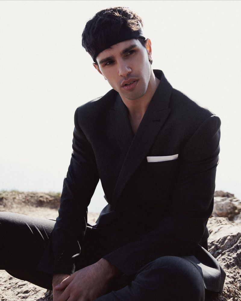 Portrait of a young adult male wearing a black suit and headband, seated outdoors against a bright, out-of-focus background.