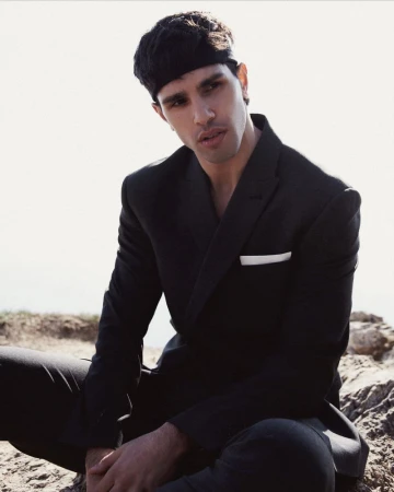 Portrait of a young adult male wearing a black suit and headband, seated outdoors against a bright, out-of-focus background.