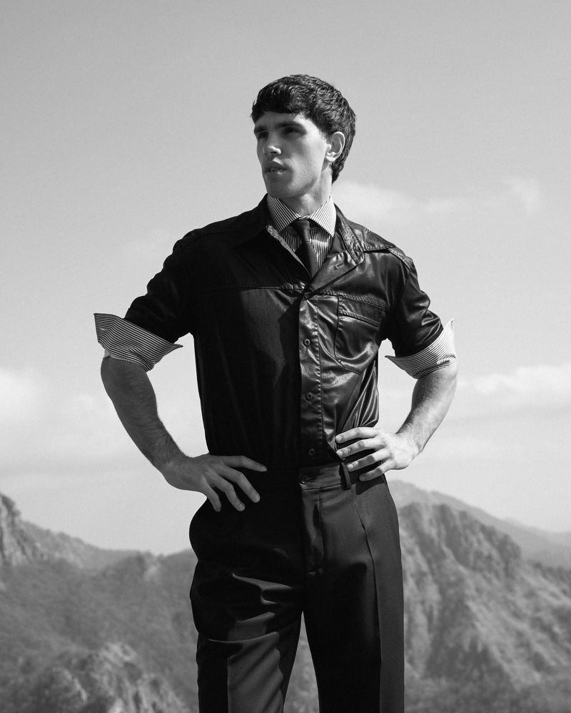 Black and white photo of Denis Jovanovic in a dark collared shirt and trousers standing with hands on hips against mountains.