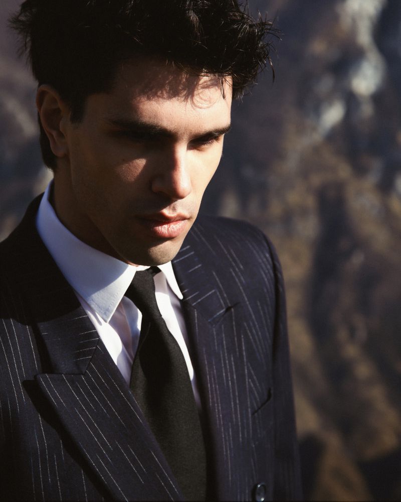 Model Denis Jovanovic wearing a dark pinstripe suit jacket, white collared shirt, and black tie against a blurred mountain backdrop.