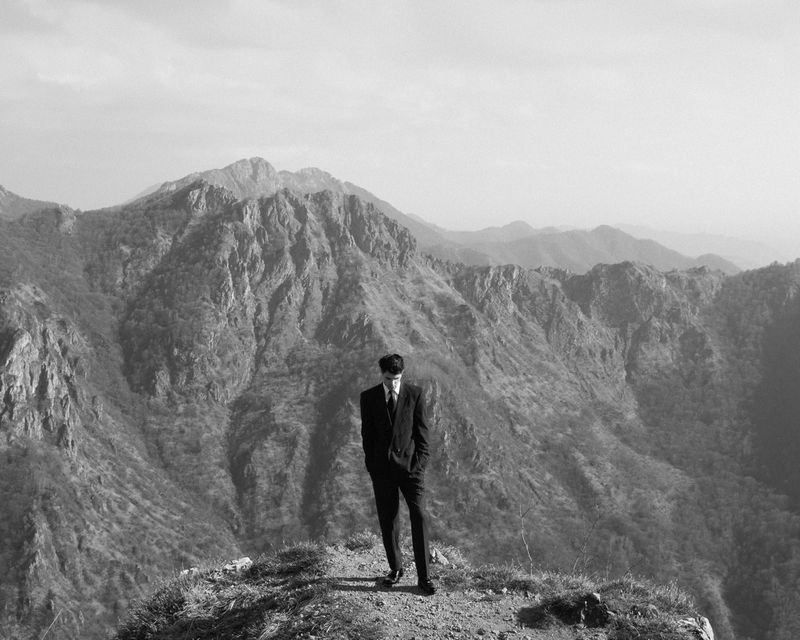 Black and white photo of a man in a dark suit standing on a mountain peak overlooking rugged ridges.