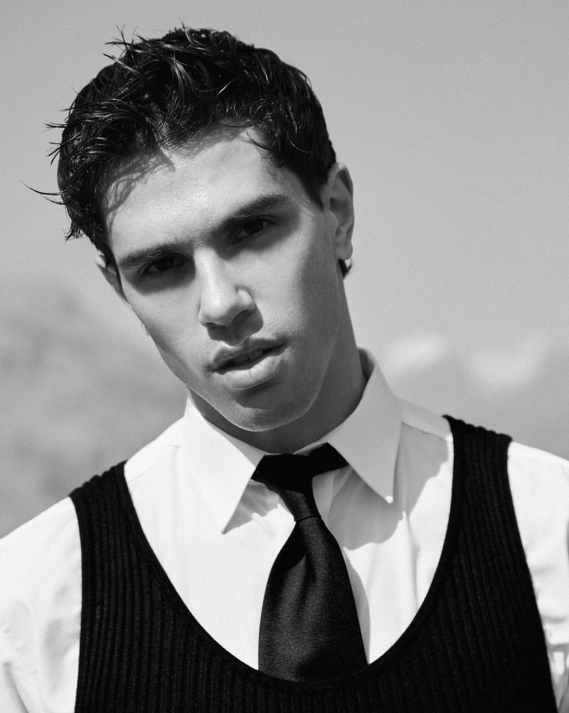 Black and white portrait of a young man in a white shirt, black vest, and tie looking at the camera against a bright sky background.