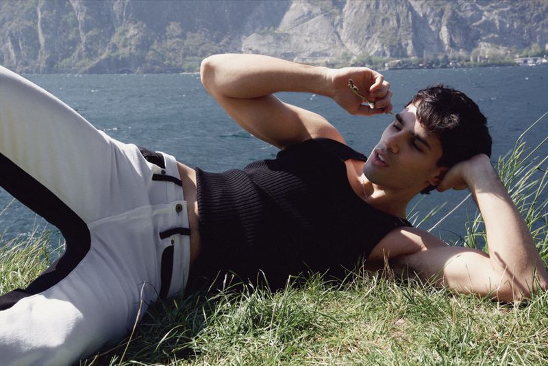 Adult male model lounges on grassy hillside near water, wearing black top and white pants with mountain background, shot at eye level.