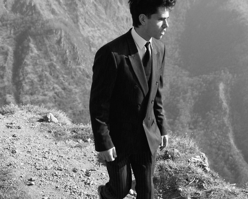 Man in a dark pin-stripe suit stands on a grassy mountain peak against a backdrop of a vast mountain range in monochrome.