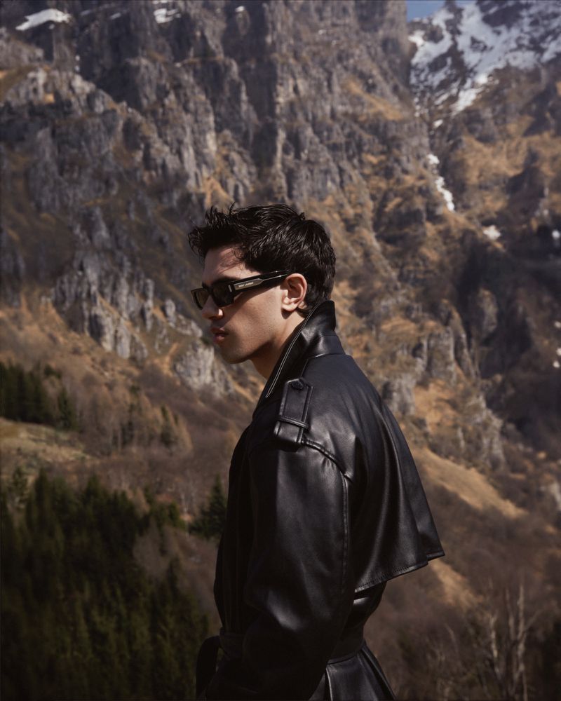 Side profile of a stylish adult male in a black leather coat and sunglasses, set against a backdrop of rugged mountains and sparse vegetation.