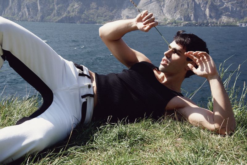 Man lying on a grassy hill near a body of water. He is wearing black tank top and white pants with black side stripes.