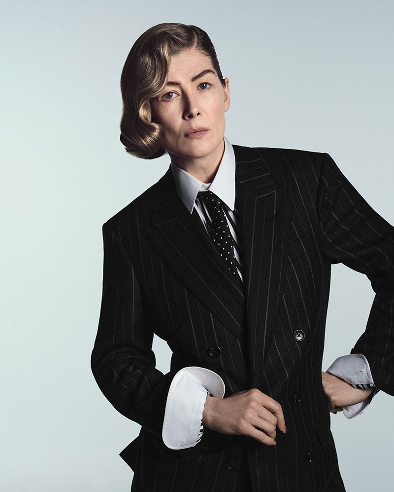 Rosamund Pike posing in a black pinstripe double-breasted suit and white collared shirt with a polka-dot tie.