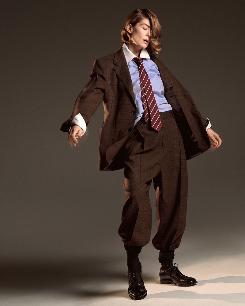 Rosamund Pike in an oversized brown tailored suit, blue collared shirt, and striped tie posing against a neutral studio backdrop.