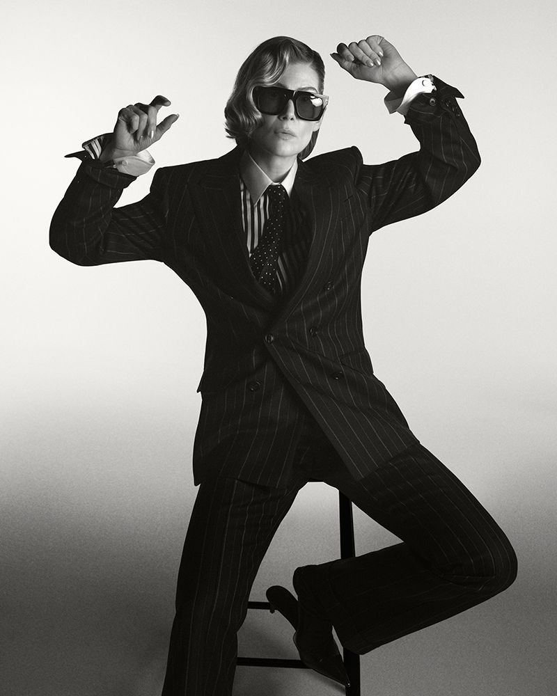 Rosamund Pike wearing a pinstripe suit and sunglasses, seated on a stool against a white seamless backdrop.