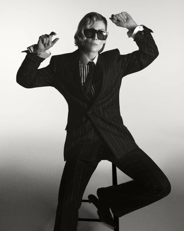 Rosamund Pike wearing a pinstripe suit and sunglasses, seated on a stool against a white seamless backdrop.