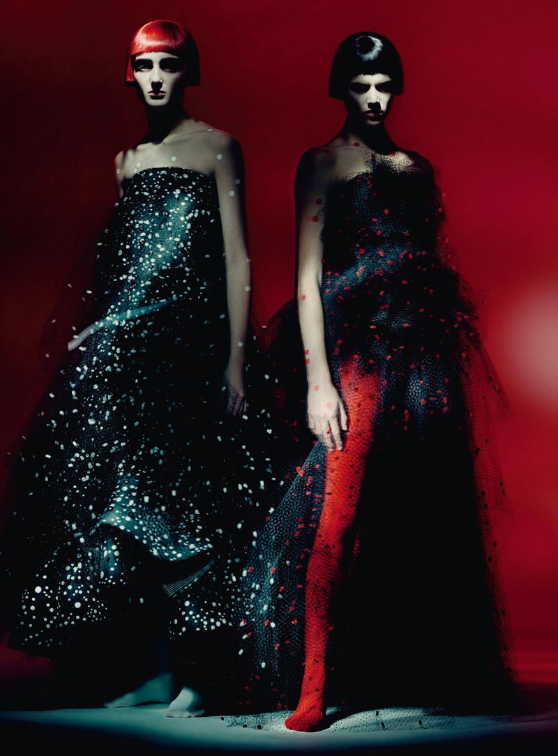 Two female models in dark textured couture gowns standing against a dramatic red and black background.