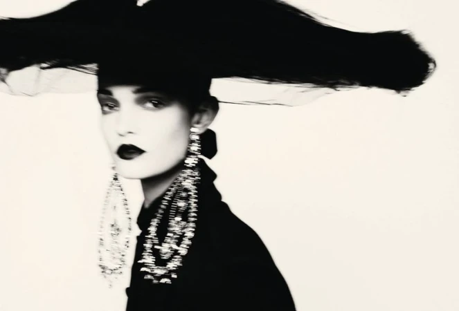 Monochrome portrait of a model wearing a wide black headpiece and sparkling crystal chandelier earrings against a white background.