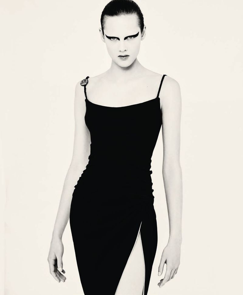Model posing in a black slip dress with a high thigh slit and dramatic winged graphic eye makeup against a white backdrop.