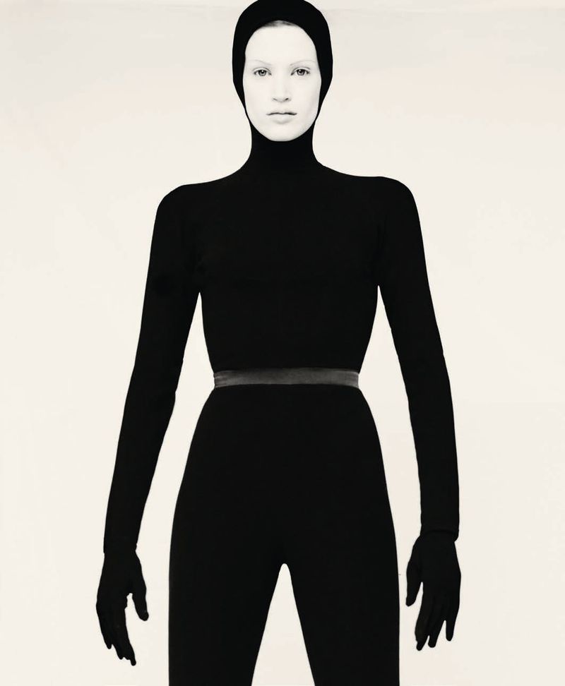Model wearing a form-fitting black hooded bodysuit with attached gloves against a plain light studio backdrop.