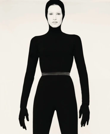 Model wearing a form-fitting black hooded bodysuit with attached gloves against a plain light studio backdrop.