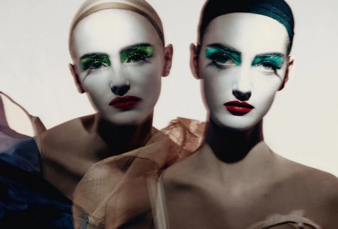 Two female models with dramatic white face makeup, green glitter eyeshadow, and red lipstick against a plain light background.