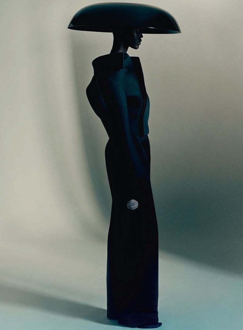 Profile of a model in a sculptural black haute couture garment and oversized dome headpiece against a grey studio backdrop.