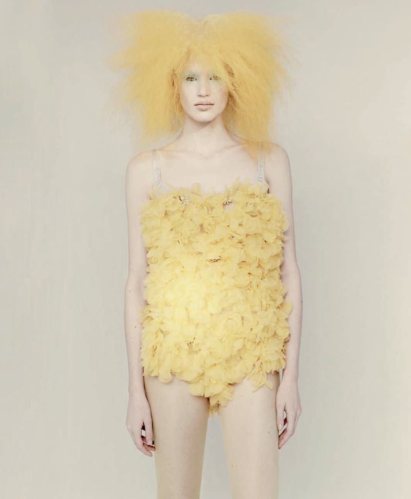 Fashion model with voluminous yellow teased hair wearing a yellow ruffled minidress against a pale seamless backdrop.
