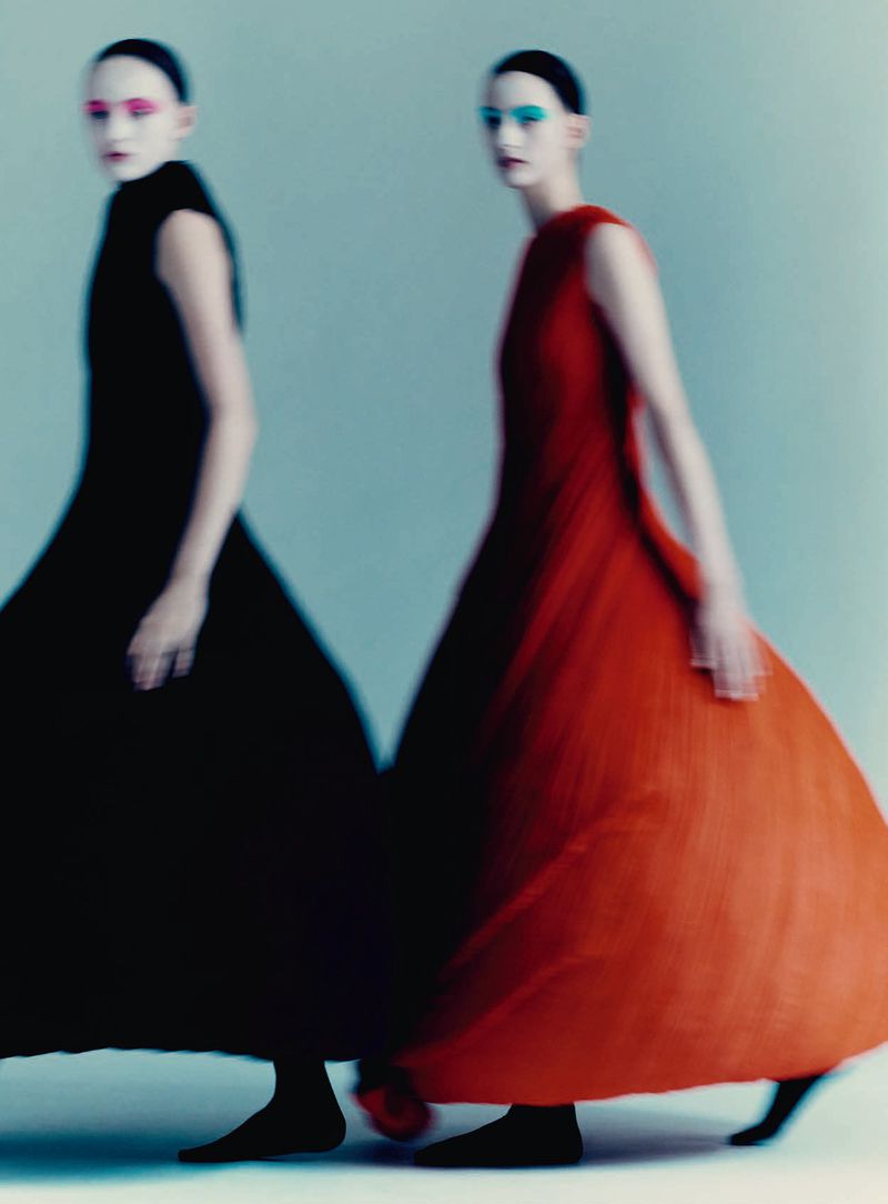 Two models in motion wearing long black and orange dresses against a plain studio backdrop