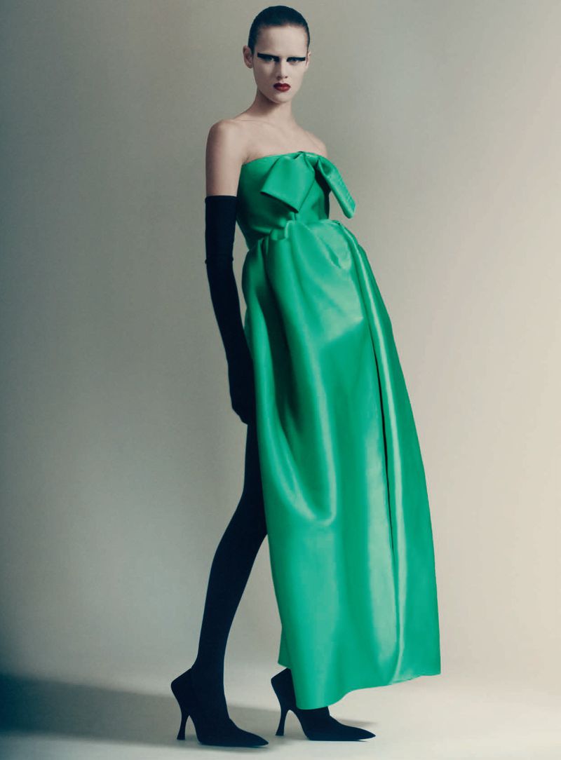 Model posing in an emerald green strapless gown paired with long black gloves and stiletto pumps against a neutral studio backdrop.