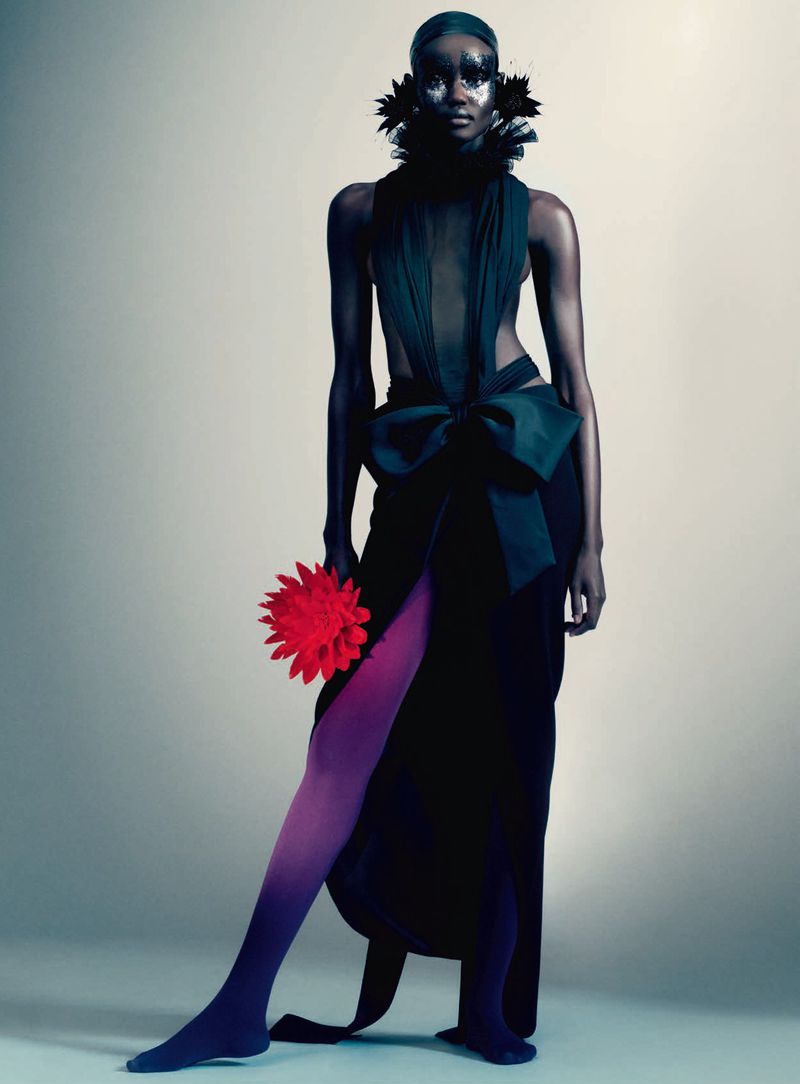 Model in dark gown with large bow holding a red flower against a neutral gradient background