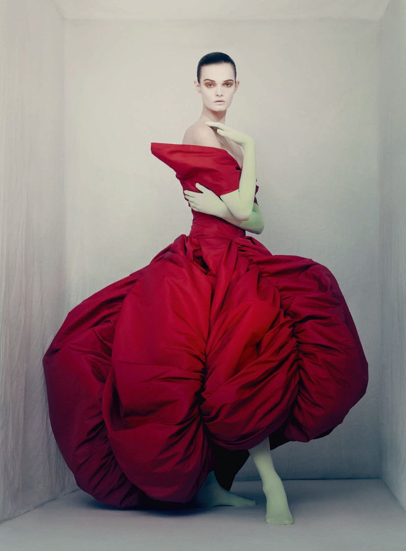 Model Lulu Tenney posed in a voluminous sculptural red haute couture gown and pale green gloves inside a neutral studio corner.