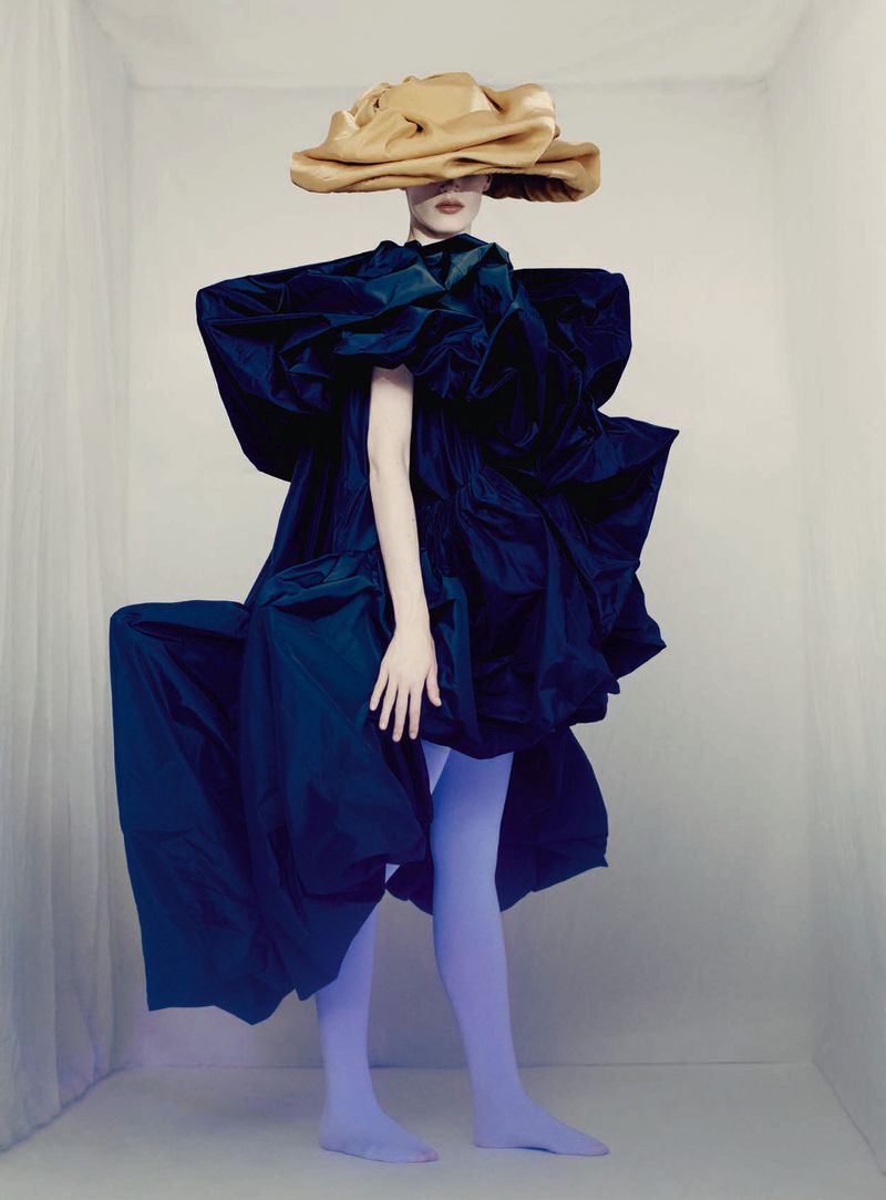 Model wearing an oversized camel headpiece, voluminous dark blue couture gown, and lavender tights against a pale studio backdrop.