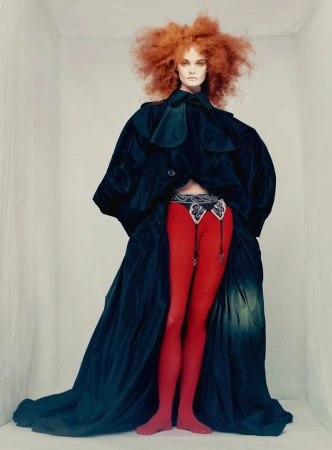 Model with voluminous red hair wearing a dark oversized coat and red leggings against a white background