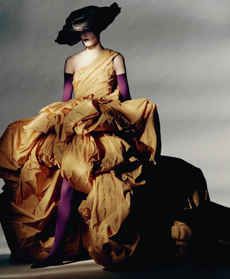 Model wearing a voluminous yellow taffeta gown, purple gloves, and an oversized black hat in a studio editorial setting.