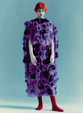 Model with orange hair wearing a purple floral cape and red socks against a pale blue background