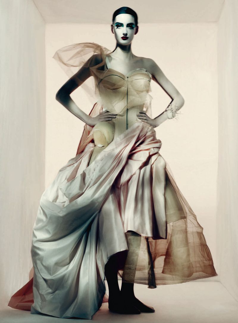Model wearing a structured sheer corset gown with voluminous draped skirts standing against a pale seamless studio background.