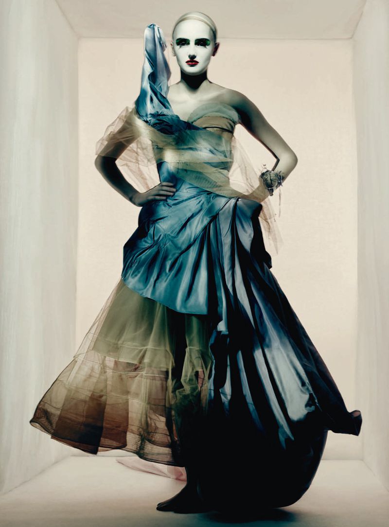 Model in a sculptural blue and green haute couture gown with avant-garde makeup against a plain background.