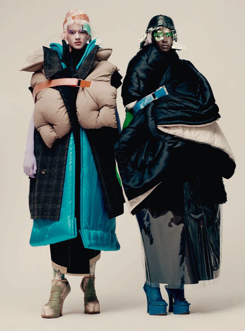 Two fashion models posing in voluminous layered puffer coats and platform footwear against a plain neutral backdrop.