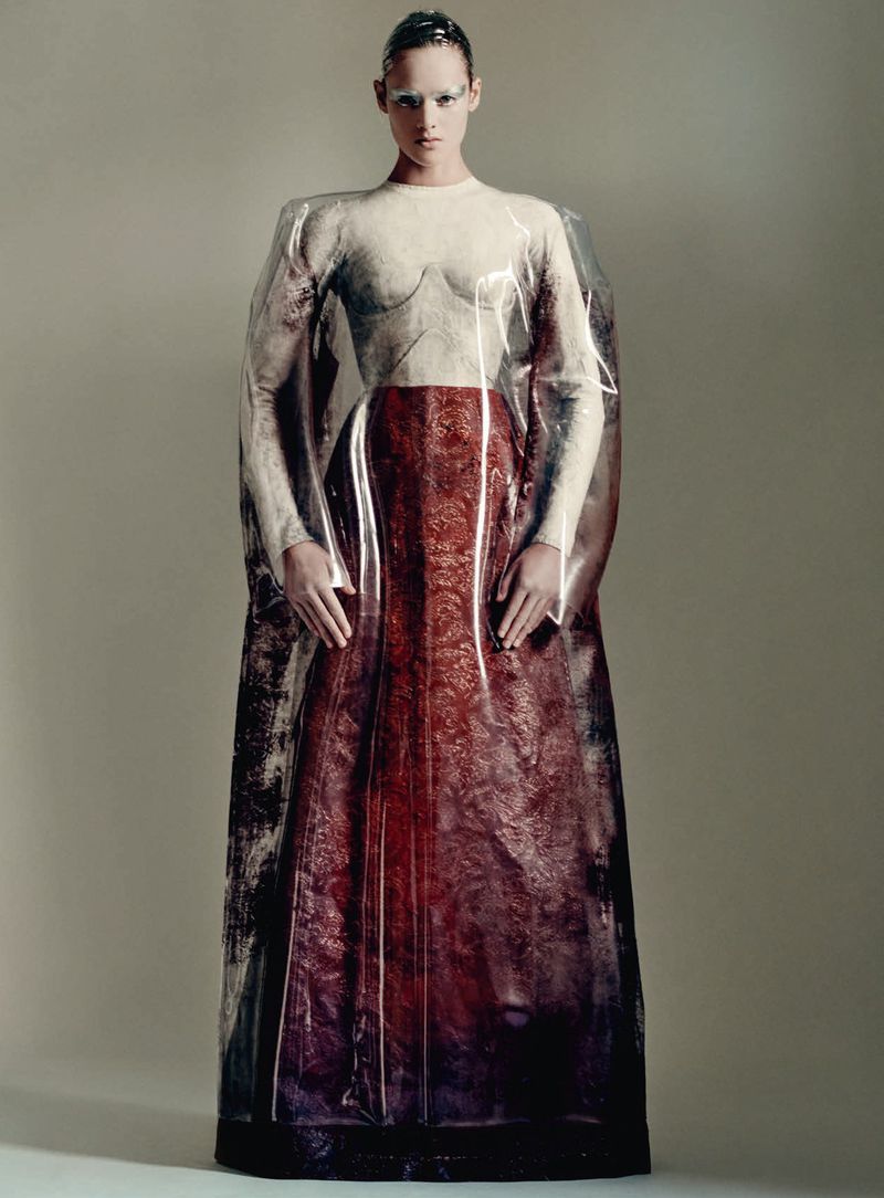 Model posing in a structured white and burgundy gown encased in a clear plastic shroud against a neutral studio backdrop.
