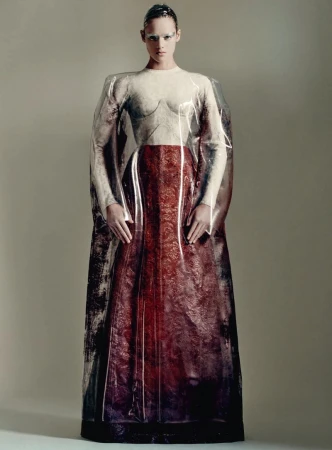 Model posing in a structured white and burgundy gown encased in a clear plastic shroud against a neutral studio backdrop.